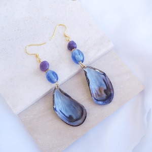Peut inclure: Une paire de boucles d'oreilles pendantes avec des crochets dorés, chacune ornée d'une petite perle violette, d'une perle bleue translucide et d'un grand pendentif en coquille de moule bleu foncé irisé au fini lisse et poli.
