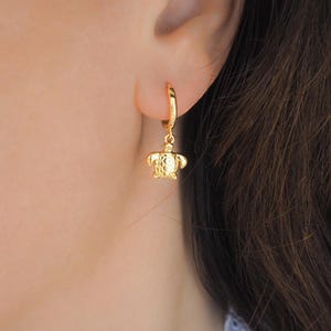 Peut inclure: Boucle d'oreille dorée avec un petit pendentif en forme de tortue. Le pendentif tortue présente des détails texturés. La boucle d'oreille est portée sur une oreille, l'accent étant mis sur le bijou.