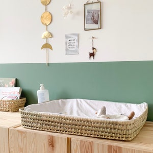 Könnte beinhalten: Ein natürlicher geflochtener Korb mit einem weißen Spannbettlaken und einem Stofftier darin. Der Korb steht auf einer Kommode aus Holz mit einer grünen Wand dahinter. An der Wand über der Kommode befinden sich Dekorationen, darunter ein goldener Mond- und Sonnen-Mobile, ein gerahmtes Foto und eine Karte mit der Aufschrift "Ich liebe meine Mama sehr".