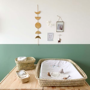 May include: A light green and white nursery with a wicker changing mat, three wicker baskets, and a gold moon and star wall hanging. The wall hanging has the text "I love my mom, love lots, love you, and lets".