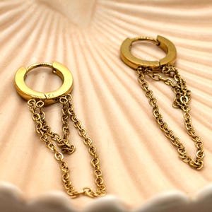 May include: A pair of gold-toned hoop earrings, each featuring two delicate dangling chains attached to the bottom of the hoop. The earrings have a smooth, polished finish and a hinged closure mechanism, creating a layered and textured appearance.
