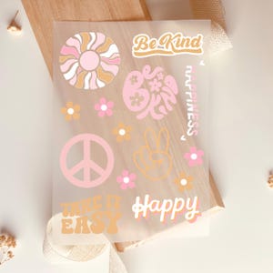Può includere: Un foglio trasparente con adesivi in stile retrò con un simbolo di pace, fiori e le parole "Be Kind", "Take it Easy" e "Happy". Gli adesivi sono in tonalità di rosa, arancione e beige.