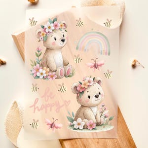 Può includere: Un foglio di adesivi con due orsetti con corone floreali, circondati da fiori, farfalle, api e un arcobaleno. La frase "be happy" è scritta in corsivo. La palette di colori include rosa, verde e giallo tenui.