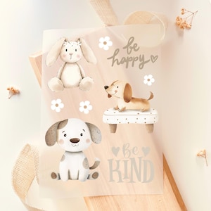 Peut inclure: Un sac cadeau transparent avec des illustrations d'un lapin et de deux chiens, accompagnées des phrases « soyez heureux » et « soyez gentil ». Le lapin porte une couronne florale, et les chiens sont de style dessin animé. Des fleurs blanches ornent les illustrations.