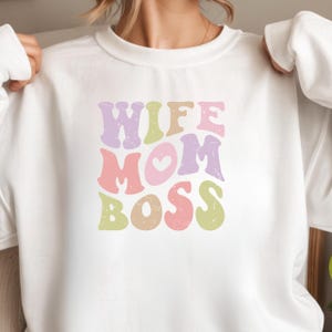 Könnte beinhalten: Ein weißes Sweatshirt mit Rundhalsausschnitt und einem Grafikdruck mit dem Text WIFE MOM BOSS in einer wellenförmigen, retro-inspirierten Schriftart. Die Buchstaben sind in Pastelltönen wie Lavendel, Hellgrün, Beige und Rosa gehalten.