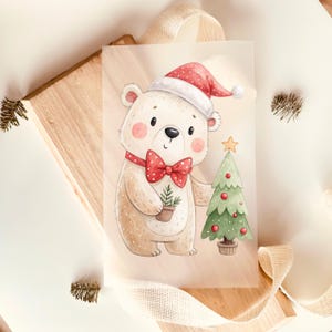 Könnte beinhalten: Eine rechteckige Grußkarte mit einer Aquarellillustration eines hellbraunen Bären, der eine rote Weihnachtsmannmütze und eine rote Fliege mit weißen Punkten trägt. Der Bär hält eine kleine Topfpflanze neben einem geschmückten grünen Weihnachtsbaum.