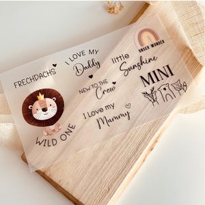 Könnte beinhalten: Eine transparente Aufkleberfolie mit einer Löwenillustration, den Worten "Ich liebe meinen Papa", "Neu im Team", "Ich liebe meine Mama", "Frechdachs", "Wild One", "Kleiner Sonnenschein", "Mini" und "Unser Wunder".