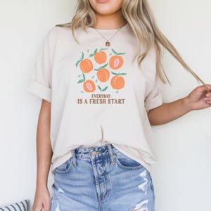 Può includere: Una maglietta beige con una grafica di pesche e il testo "EVERYDAY IS A FRESH START".