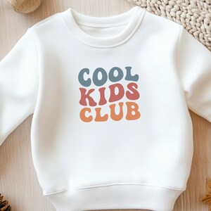 Könnte beinhalten: Weißes Sweatshirt mit Rundhalsausschnitt und dem Text "COOL KIDS CLUB" in blauen, roten und orangefarbenen Blockbuchstaben. Das Sweatshirt ist aus weichem Material und für Kinder konzipiert.