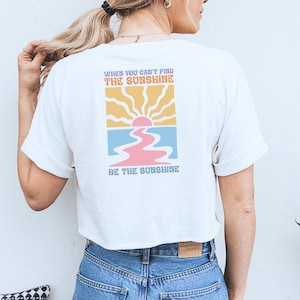 Op de afbeelding: Wit T-shirt met een afbeelding van een zonsondergang boven een roze en blauwe rivier. De tekst "When you can't find the sunshine be the sunshine" staat op het shirt gedrukt.