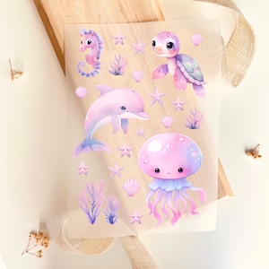 Könnte beinhalten: Ein Stickerbogen mit Aquarellillustrationen von Meereslebewesen. Die Aufkleber zeigen ein Seepferdchen, eine Schildkröte, einen Delfin und eine Qualle in Rosa- und Lilatönen. Auch Seesterne und Korallen sind enthalten.