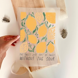 Può includere: Una stampa traslucida con limoni gialli, foglie verdi e bacche rosa. Il testo "THE SWEET JUST ISN'T AS SWEET WITHOUT THE SOUR" è stampato in basso. La stampa è su una tavola di legno.