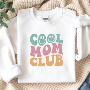 Könnte beinhalten: Weißes Sweatshirt mit dem Text "COOL MOM CLUB" in Retro-Schrift. Das Wort "COOL" ist in Türkis gehalten, mit Smiley-Grafiken. "MOM" ist in Rosa und "CLUB" in Orange. Das Sweatshirt ist aus einem weichen Material.