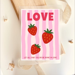 Könnte beinhalten: Eine rechteckige Grußkarte mit einem Hintergrund aus hellrosa und weißen vertikalen Wellenstreifen. Das Wort LOVE ist oben in großen roten Buchstaben gedruckt. Drei rote Erdbeeren mit grünen Blättern sind mittig platziert. Der Text LET ALL THAT YOU DO BE DONE IN LOVE steht unten in kleinen roten Buchstaben.
