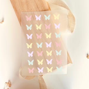 Può includere: Un foglio di materiale trasparente decorato con file di adesivi a farfalla dai colori pastello. Le farfalle sono in tonalità di rosa, giallo, blu e viola. Il foglio poggia su una superficie di legno con un nastro beige.