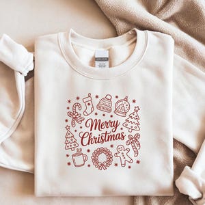 Könnte beinhalten: Cremefarbenes Sweatshirt mit roter Grafik mit der Aufschrift "Merry Christmas". Die Grafik enthält weihnachtliche Symbole wie Strumpf, Zuckerstange, Weihnachtsbaum und Lebkuchenmann.