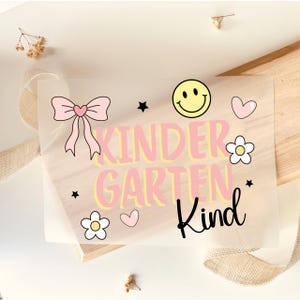 Könnte beinhalten: Ein durchscheinendes Blatt mit dem Text KINDERGARTEN Kind in rosa und gelben Buchstaben. Das Design enthält eine rosa Schleife, ein gelbes Smiley-Gesicht, zwei weiße Blumen mit gelber Mitte, zwei kleine rosa Herzen und zwei schwarze Sterne.