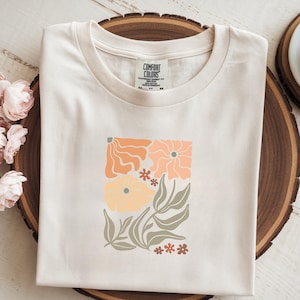 Könnte beinhalten: Cremefarbenes T-Shirt mit einem Blumendesign in Orange-, Pfirsich- und Gelbtönen. Die Grafik zeigt stilisierte Blumen und Blätter. Das Etikett lautet "COMFORT COLORS".