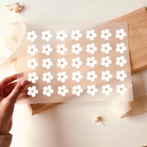 Könnte beinhalten: Ein Bogen mit weißen Gänseblümchen-Aufklebern mit braunen Zentren. Die Aufkleber befinden sich auf einem durchscheinenden Bogen, der von einer Hand gehalten wird. Der Hintergrund umfasst ein Holzbrett, Jutegewebe und Trockenblumen.