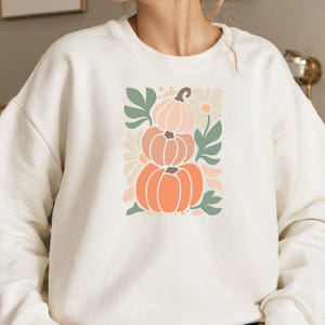 Könnte beinhalten: Cremefarbenes Sweatshirt mit einem gestapelten Kürbisdesign in Orange- und Pfirsichtönen, ergänzt durch grüne Blätter und weiße Gänseblümchen-Akzente. Die Grafik befindet sich mittig auf der Vorderseite des langärmeligen Oberteils.