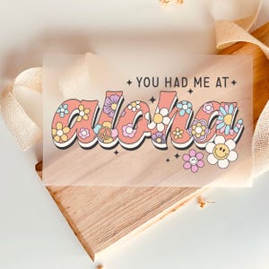 Könnte beinhalten: Ein transparenter Aufkleber mit dem Text "You had me at Aloha" in rosa und schwarzer Schrift. Das Wort Aloha ist mit bunten Blumen und Friedenszeichen verziert.