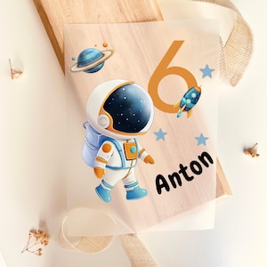 May include: A transparent sheet featuring a cartoon astronaut in a white and orange spacesuit, with a blue backpack and boots. The design includes a planet, rocket, stars, the number 6, and the name "Anton" in black.