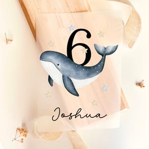 Könnte beinhalten: Transparente Acryltafel mit einer Illustration eines blauen Wals, der Zahl 6 und dem Namen "Joshua". Der Wal hat weiße Details und Sterne. Der Hintergrund ist hellpfirsichfarben. Maße in Zentimetern.