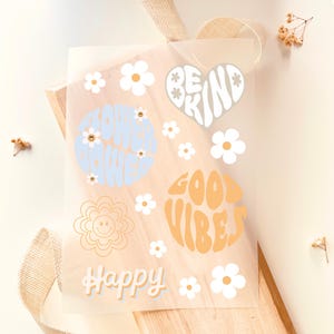 Può includere: Un foglio di adesivi trasparente con vari disegni floreali e affermazioni positive. Il foglio include le frasi "Flower Power", "Be Kind", "Good Vibes" e "Happy", insieme a diverse illustrazioni di margherite in colori pastello.