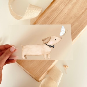 Könnte beinhalten: Hellbraune Karte mit einem Cartoon-Dackel mit schwarzer Fliege, der einen hellblauen Schmetterling ansieht. Der Hund hat rosa Wangen und braune Ohren. Die Karte wird von einer Hand gehalten, mit Holz und Band im Hintergrund.