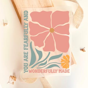 Könnte beinhalten: Ein rechteckiger Druck mit einer großen rosa Blume mit gelber Mitte, blaugrünen Blättern und einer kleineren gelben Blume. Der Text "YOU ARE FEARFULLY AND WONDERFULLY MADE" ist in Blaugrün und Rosa vertikal und horizontal angeordnet.