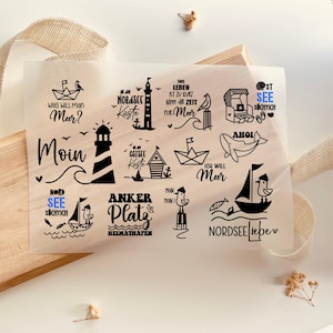 Op de afbeelding: Een transparante folie met zwarte nautische illustraties en tekst. Ontwerpen omvatten een vuurtoren, zeilboot, walvis en zinnen als "Moin" en "Nordsee Liebe". De folie ligt op een houten oppervlak.