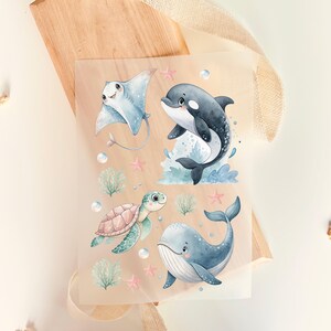 Könnte beinhalten: Ein Blatt Papier mit Aquarellillustrationen von Meereslebewesen. Das Design umfasst einen Mantarochen, einen Orca, eine Meeresschildkröte und einen Wal sowie Seesterne und Blasen. Die Farbpalette ist weich, mit Blau-, Grün- und Rosatönen.