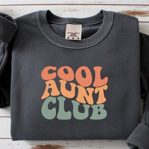 Könnte beinhalten: Dunkelgraues Sweatshirt mit dem Schriftzug "COOL AUNT CLUB" in orangefarbenen und grünen Retro-Lettern. Das Sweatshirt hat einen Rundhalsausschnitt und lange Ärmel. Der Hintergrund ist ein helles Holzmaserungsmuster.