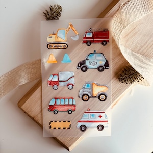 Könnte beinhalten: Ein Bogen mit bunten Aufklebern, die verschiedene Fahrzeuge zeigen, darunter einen gelben Bagger, ein rotes Feuerwehrauto, ein blaues Polizeiauto, einen grauen Lieferwagen, einen grauen Kipplaster, einen roten Bus und einen weißen Krankenwagen. Die Aufkleber befinden sich auf einem transparenten Hintergrund.