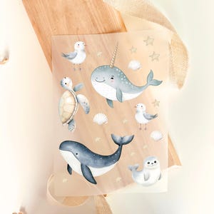 Op de afbeelding: Een vel stickers met aquarel illustraties van zeedieren. De stickers bevatten een narwal, walvis, schildpad, zeehond en zeemeeuwen, samen met schelpen en sterren. Het kleurenpalet is zacht, met blauw, wit en beige.