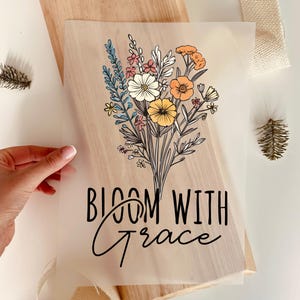 Könnte beinhalten: Ein Blumendesign mit den Worten "Bloom With Grace" in Schwarz gedruckt. Der Strauß zeigt weiße, gelbe, orangefarbene und rosa Blumen mit grünem und blauem Laub. Das Design befindet sich auf einem durchscheinenden Blatt.