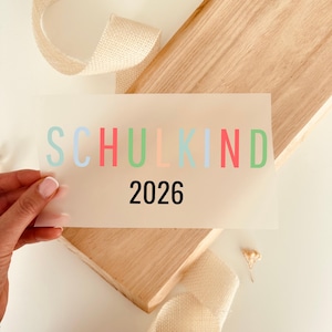Könnte beinhalten: Ein rechteckiger, halbtransparenter Transferbogen mit dem Wort SCHULKIND in abwechselnd pastellblauen, roten und grünen Buchstaben, wobei das Jahr 2026 in fetter, schwarzer serifenloser Schrift unter dem Text auf einer hellen Holzoberfläche steht.