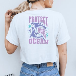 Könnte beinhalten: Weißes, bauchfreies T-Shirt mit einem violetten Grafikdruck von zwei Walen, einem Seepferdchen, einer Schildkröte und dem Text "PROTECT OUR OCEAN".