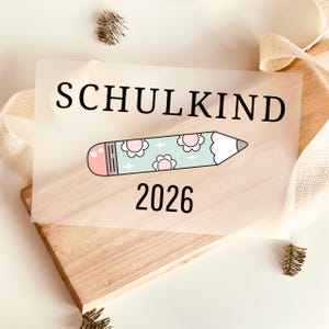 Peut inclure: Une feuille rectangulaire translucide portant le texte noir SCHULKIND au-dessus d'un dessin de crayon bleu clair avec une gomme rose et des motifs de fleurs blanches, suivi du texte noir 2026, posée sur une planche en bois clair.