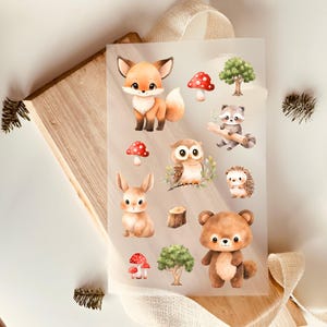 Könnte beinhalten: Eine Blatt mit transparenten Vinyl-Aufklebern mit Waldtieren, darunter ein Fuchs, eine Eule, ein Kaninchen, ein Waschbär, ein Igel und ein Bär. Die Aufkleber sind im Aquarell-Stil mit braunen, orangen und grauen Tönen illustriert.