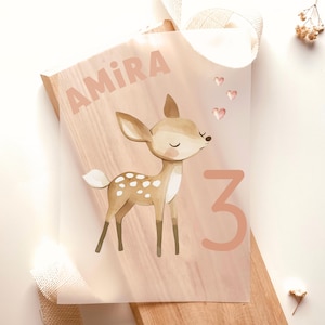 Könnte beinhalten: Personalisierter Geburtstagsdruck mit einer Illustration eines Rehs mit weißen Flecken, dem Namen "AMIRA", der Zahl "3" und drei rosa Herzen. Das Design befindet sich auf einer durchscheinenden Folie über einer Holzoberfläche.