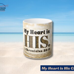 May include: White ceramic mug with a gold glitter background and black text that reads "My Heart is HIS. 1 Chronicles 22:19". The mug is sitting on a sandy beach with a blue ocean in the background.