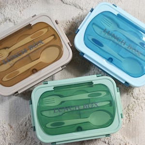 Könnte beinhalten: Drei Lunchboxen mit integriertem Besteck. Die Lunchboxen sind beige, blau und grün. Jede Lunchbox enthält eine Gabel, ein Messer und einen Löffel.