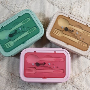 Könnte beinhalten: Drei Lunchboxen mit integrierten Bestecken. Die Lunchboxen sind grün, braun und rosa. Jede Lunchbox enthält einen Löffel und eine Gabel. Die Deckel sind transparent und zeigen das Besteck. Die Lunchboxen tragen den Text "life nutritionist".