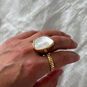Puede incluir: Un anillo dorado con una gran incrustaci&oacute;n irregular de n&aacute;car blanco liso y un segundo anillo dorado con una textura trenzada similar a una cuerda. Ambos anillos tienen un acabado met&aacute;lico pulido y se llevan en los dedos de una mano.