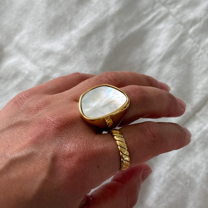 Puede incluir: Un anillo dorado con una gran incrustaci&oacute;n irregular de n&aacute;car blanco liso y un segundo anillo dorado con una textura trenzada similar a una cuerda. Ambos anillos tienen un acabado met&aacute;lico pulido y se llevan en los dedos de una mano.