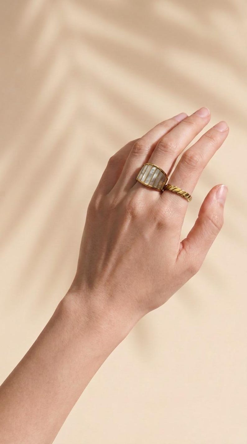 May include: A gold-toned ring featuring a wide, curved band inlaid with vertical, rectangular segments of iridescent white mother-of-pearl, accompanied by a second, thinner gold-toned ring with a twisted, rope-like texture on the adjacent finger.