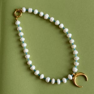 Puede incluir: Un delicado collar con perlas blancas ensartadas en un cordón azul. El collar tiene un cierre dorado y un colgante de media luna. Las perlas tienen forma irregular, lo que añade un toque único a la joya.