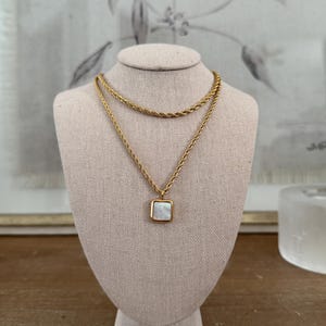 May include: A gold chain necklace with a square pendant featuring a white mother-of-pearl inlay. The necklace is layered with a second, thinner gold chain.