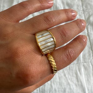 May include: A wide gold-tone ring featuring vertical inlays of iridescent white mother-of-pearl segments. The band is connected to a secondary, thinner gold-tone ring with a twisted, rope-like texture that sits lower on the finger.